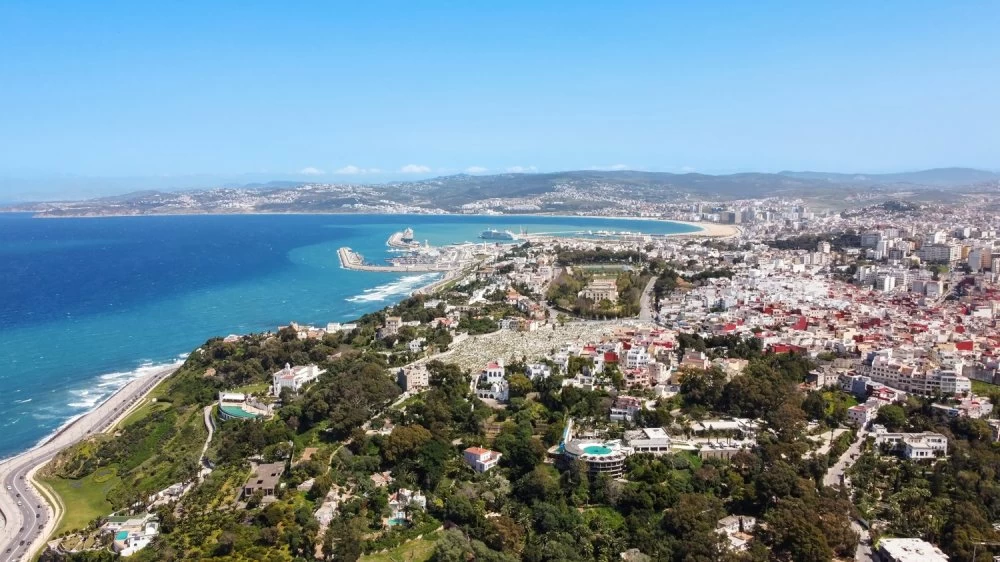 Aerial drone view of Tangier, Morocco. Strait of Gibraltar coastline with multiple residential buildings and lush greenery located on rocky hills, sea port and beach in the distance